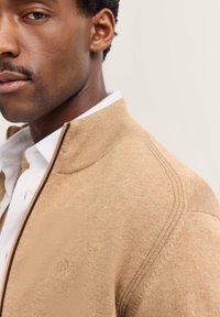 Maglione beige con zip, colletto alto, texture a coste e un motivo sottile. Presenta un piccolo logo sulla parte sinistra del petto.