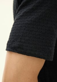 Czarny, krótki sweter z dzianiny z teksturowanym wzorem i lekko dopasowanym krojem, odsłaniający część jasnobarwnego ramienia poniżej.