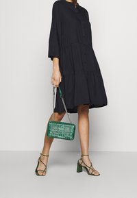 Svart, långärmad knapprad klänning med tierad design. Grön, texturerad handväska med kedjehandtag och matchande gröna klackar med remmar.