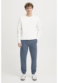 Sudadera blanca con cuello redondo, combinada con pantalones de chándal azules. Los pantalones tienen un corte holgado. Zapatillas con rayas negras y blancas.