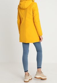 Gul huva jacka med en strömlinjeformad design, kombinerad med blå tajta jeans och beige sneakers med en kraftig vit sula.