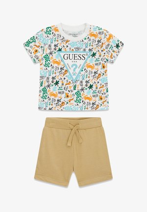 Conjunto para niño pequeño que incluye una camiseta blanca de manga corta con gráficos coloridos de temática playera y el logo "GUESS", combinada con pantalones cortos beige lisos con cordón.