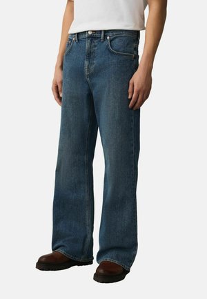 Mann, der blaue Jeans mit weitem Bein, ein weißes T-Shirt und dunkelbraune Stiefel trägt, vor einem schlichten hellen Hintergrund steht.