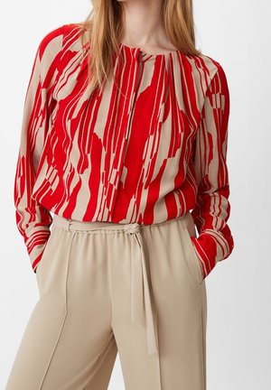 Femme portant un chemisier rayé abstrait rouge et beige, rentré dans un pantalon beige taille haute avec une ceinture nouée en tissu.