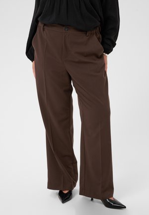 Personne portant un pantalon marron ample et des talons noirs à bout pointu, debout devant un fond uni.