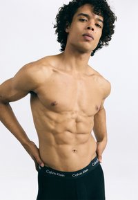 Boxerii negri Calvin Klein au un brâu elastic cu logo. Modelul își afișează torso-ul tonifiat și muscular pe un fundal simplu.