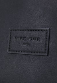 Svart läderväska med en texturerad yta och en sydd rektangulär tagg märkt "PIER ONE" och "BERLIN" i prägling.