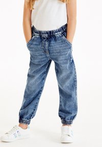 Ljust blå denimjoggers med elastisk midja, sidofickor och elastiska ärmmuddar, ihopparat med vita sneakers med blå detaljer.