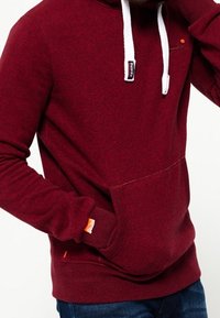 Sudadera burdeos hecha de tela texturizada con cordones blancos, bolsillo canguro y detalle de etiqueta naranja en la manga.