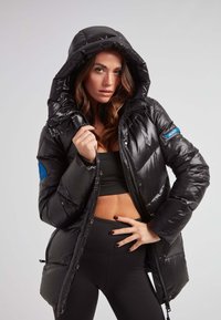 Czarna kurtka puchowa o połyskującej powierzchni, z dużym kapturem, zamkiem z przodu oraz niebieską naszywką na rękawie. Luźny krój z kieszeniami po bokach.