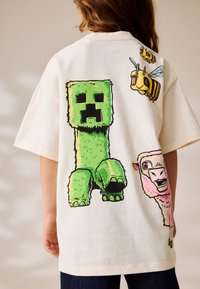 Cremefarbenes T-Shirt mit einer grünen, eckigen Figur, einer gelben Biene und einem pinken Schäfchen-Motiv auf der Rückseite. Baumwollstoff, lockere Passform.