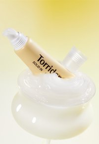 Torriden hudplejecreme i en beige tube med sort tekst, indsat i en glat, hvid cremesvulv i en klar beholder, sat mod en gul baggrund.