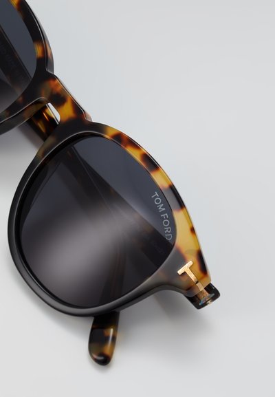 Tortoisekleurige zonnebril met zwarte accenten, grijze lenzen en een goudkleurig "T" logo op de pijp. Gedurfde, oversized vorm.