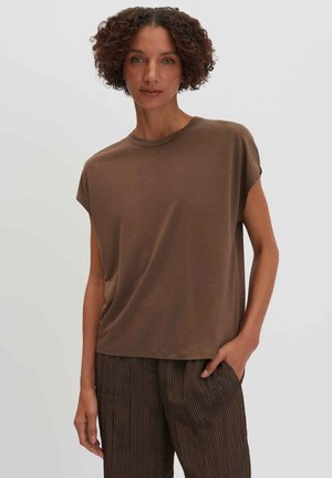 Frau mit lockigem Haar, die ein locker sitzendes braunes Kurzarmoberteil und dunkelbraune Hose mit feinen Streifen trägt, steht mit einer Hand in der Tasche.