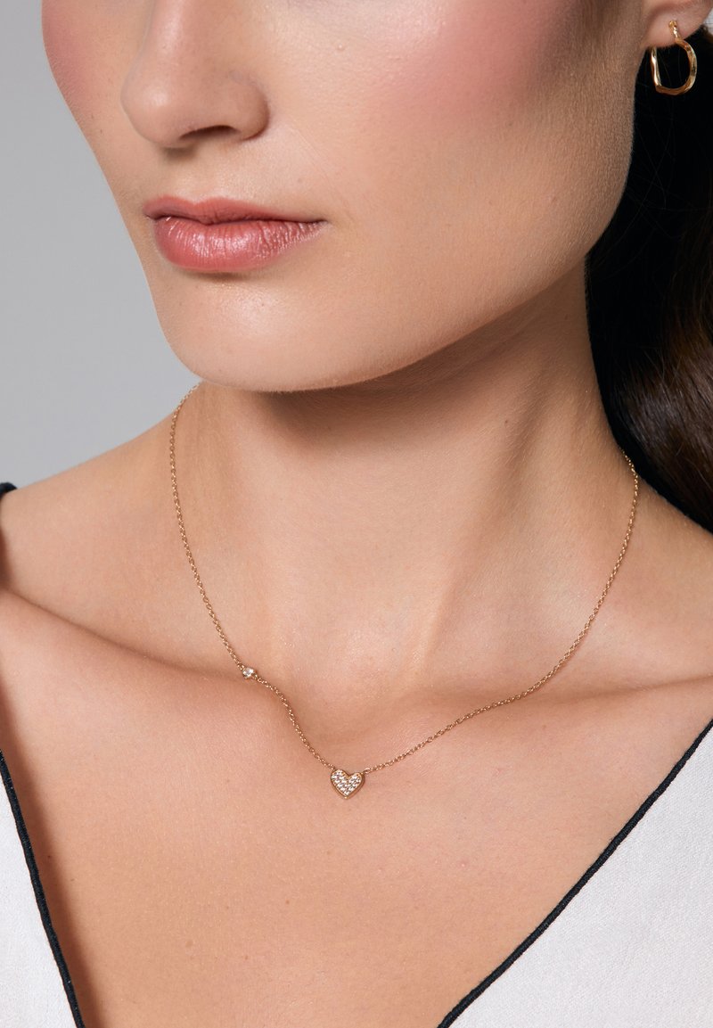 Złoty naszyjnik łańcuszkowy z serduszkiem ozdobionym małymi, przezroczystymi kamieniami, spoczywający na nagim obojczyku. Prosty i elegancki design.