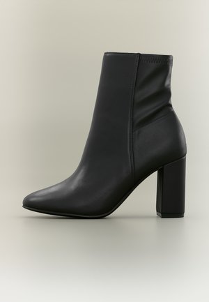 Bottine en cuir noir avec bout pointu et talon bloc haut sur un fond neutre.