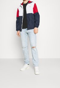 Färgblockerad vindjacka i röd, vit och marinblå, med huva och elastiska ärmslut, matchad med ljusblå distressed jeans och vita sneakers.