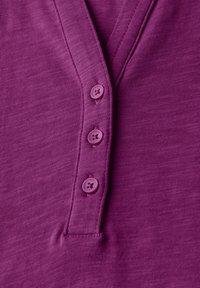 Top a maniche corte di colore viola scuro con scollatura a V e quattro bottoni abbinati. Il tessuto è testurizzato con sottili righe orizzontali.