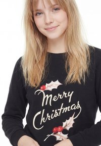 Felpa nera con la scritta "Buon Natale" e decorazioni di foglie di agrifoglio in rosso e bianco. Girocollo standard, maniche lunghe e polsini a costine.