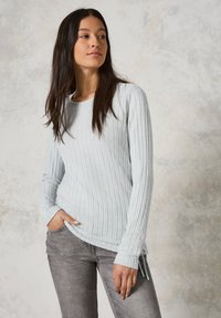 Haut turquoise à rayures, à manches longues, avec un col rond et des fentes sur les côtés, associé à un jean gris. Le tissu semble doux avec une texture subtile.