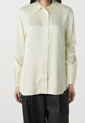 Femme portant une chemise crème à manches longues boutonnée avec un col pointu et un pantalon noir large sur un fond uni.