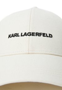 Valge puuvillane beisbolimüts, millel on kõver ääreosa. Ees on tikkitud mustad tähed, mis kirjutavad "KARL LAGERFELD".