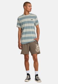 Camicia a maniche corte a righe in azzurro chiaro e crema con logo, abbinata a pantaloni corti marroni con design grafico bianco e sneaker nere.