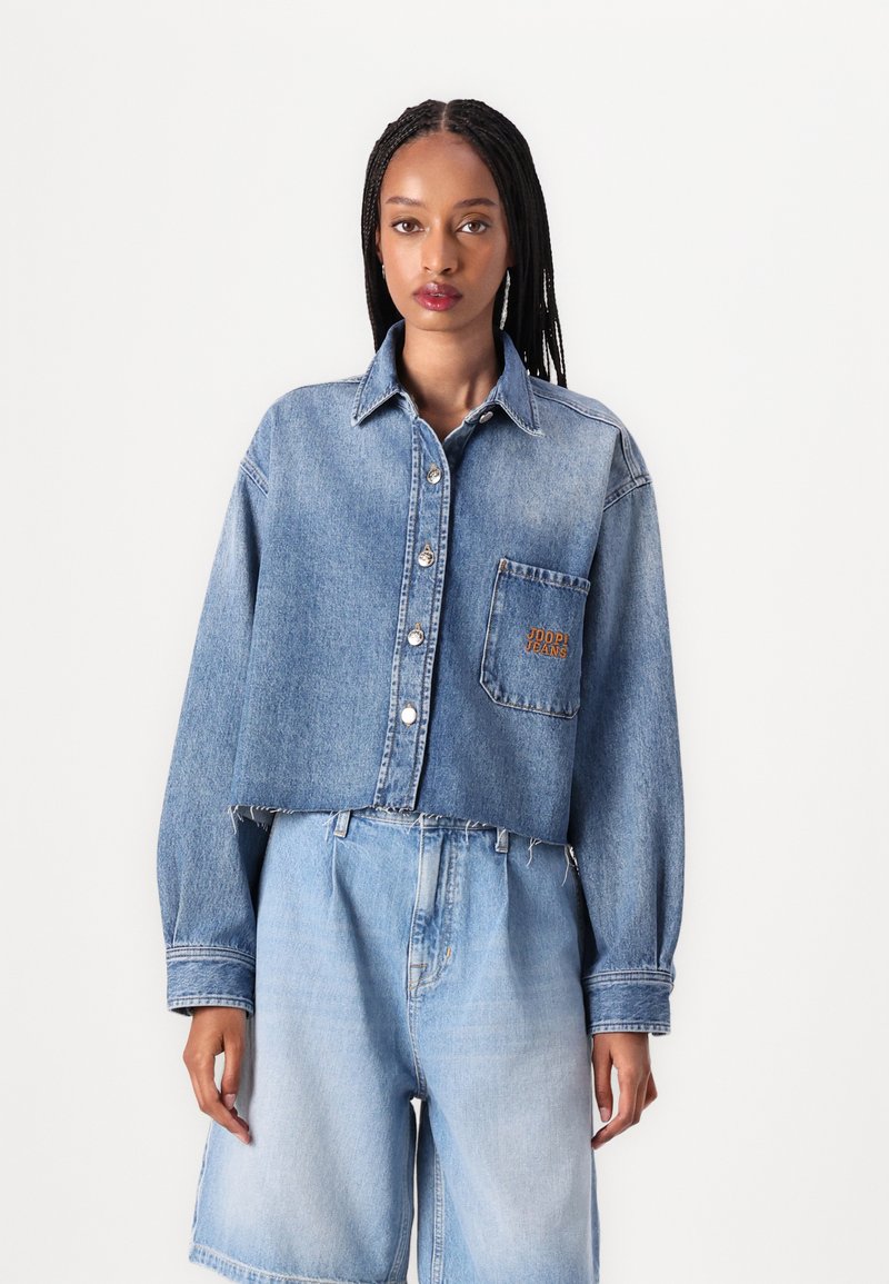 Giacca di denim tagliata in blu chiaro con maniche lunghe, chiusura con bottoni, tasca frontale con ricamo arancione "JODI JEANS" e orlo sfrangiato.
