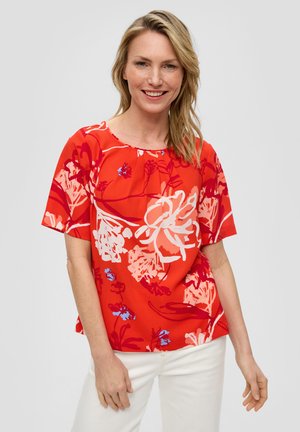 Rote Bluse mit kurzen Ärmeln, die mit weißen und blauen Blumenmustern versehen ist. Sie hat einen runden Ausschnitt und besteht aus leichtem Stoff. Kombiniert mit weißen Hosen.