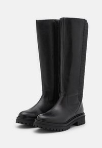 Botas altas negras de cuero liso hasta la rodilla, con cremallera lateral y detalle de malla texturizada en la parte posterior. Suela de goma gruesa con dibujo.