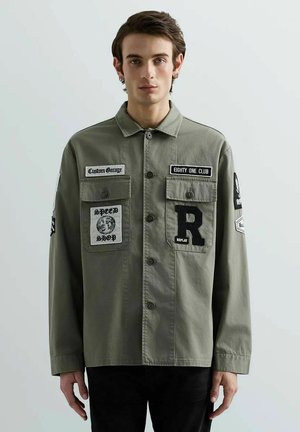 Giovane uomo con giacca verde oliva a bottoni con toppe con scritte "Custom Garage", "Eighty One Club" e "Replay" in piedi davanti a uno sfondo semplice.