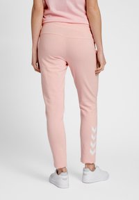 Ljusrosa sweatpants med en slät textur, elastisk midja och vitt chevronmönster på vänster ben, i kombination med vita träningssneakers.