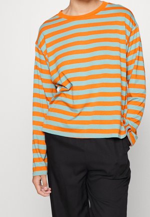 Personne portant une chemise à manches longues à rayures horizontales orange et bleu clair avec un pantalon noir, main reposant sur le côté.