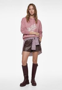 Roze hoodie met bloemenprint, bruine leren korte broek en gestreept shirt dat in de taille is gestrikt. Lange bordeauxrode rubberlaarzen maken de outfit compleet.