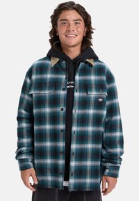 Jonge man die glimlacht, gekleed in een blauw en groen geruit overhemd over een zwarte hoodie, staand tegen een eenvoudige witte achtergrond.