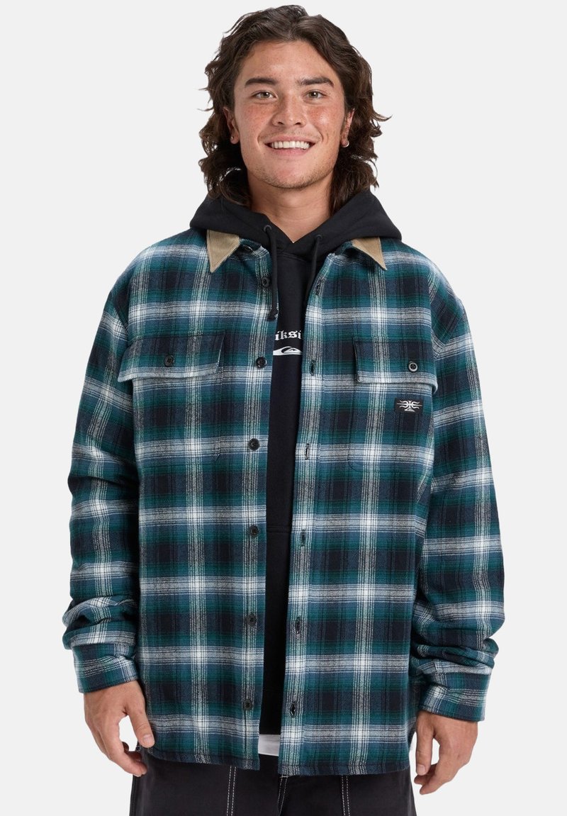 Jonge man die glimlacht, gekleed in een blauw en groen geruit overhemd over een zwarte hoodie, staand tegen een eenvoudige witte achtergrond.