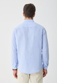 Light blue long-sleeve shirt in a soft, textured fabric. Features a pointed collar and buttoned cuffs, with a curved hem and relaxed fit.