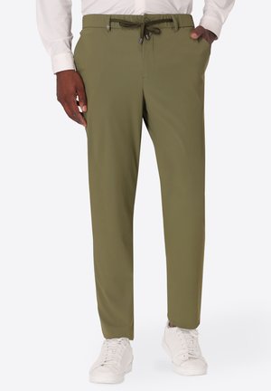 Uomo che indossa pantaloni verde oliva con coulisse, scarpe da ginnastica bianche e una camicia bianca a maniche lunghe, mani in tasca, in piedi.