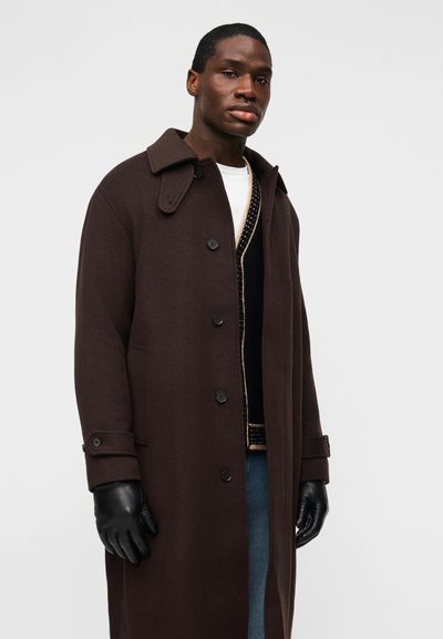 Mann trägt einen langen dunkelbraunen Knopfmantel, schwarze Lederhandschuhe, eine schwarz-beige Strickjacke, ein weißes Hemd und blaue Jeans vor einem einfarbigen Hintergrund.