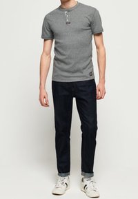 Mand iført en kortærmet grå henley-skjorte, mørke oprullede jeans og hvide sneakers, stående mod en ensfarvet lys baggrund.