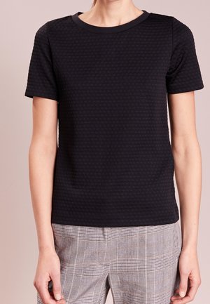 Femme portant un haut noir texturé à manches courtes associé à un pantalon gris à carreaux, debout devant un fond uni.
