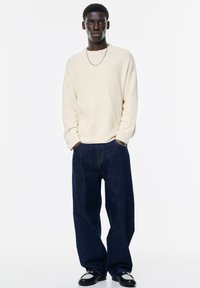 Cămașă crem, cu mânici lungi și textură cu nervuri, combinată cu blugi denim închis, cu croială lejeră și pantofi negri. Un lanț simplu din argint ca detaliu de accent.