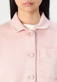 Chaqueta de mezclilla rosa claro con cuello, cierre de botón y un bolsillo en el pecho. Presenta botones plateados y detalles de costura sutiles.