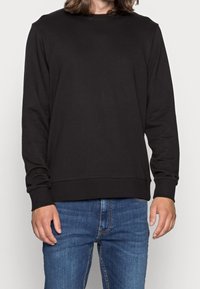 Svart långärmad sweatshirt i bomullsmaterial; har en rund halsringning och ribbade mudd med en avslappnad passform. Kombinerad med blå jeans.