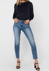 Frau in dunkelblauen Pullover, hellblauen hochgeschnittenen Skinny-Jeans und schwarzen offenen High Heels, die mit den Händen in den Taschen steht.