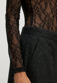 Zwarte kanten blouse met lange mouwen en bloemenpatroon, gecombineerd met hooggesneden, donkergrijze gemêleerde wollen broeken op een model.