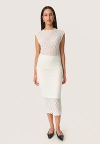 Bílá krajková šaty s upnutým střihem, rukávy ve tvaru cap, textured bodice a zig-zag lem. Spárováno s černými špičatými botami.