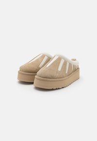 Beige zamšādas slip-on apavi ar baltām elastīgām akcentu detaļām, polsterētu apkakli un masīvu platformas zoli ar teksturētu zīmējumu.