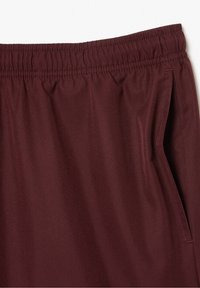 Maroon elastiska midjebyxor med subtilt diamantmönster och sidoficka på höger sida mot en vit bakgrund.