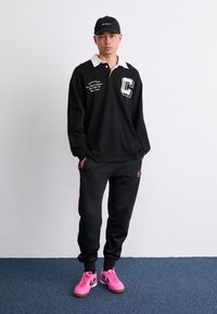 Svart langermet polo med hvit krage, med grafisk bokstav "C" og tekst. Brukt med svarte joggebukser med røde striper og rosa sko.